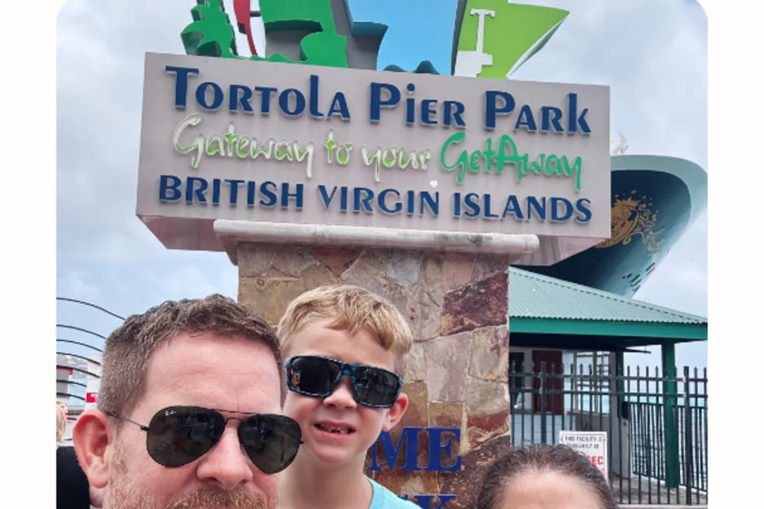 A man, a boy and a woman in sunglasses taking a selfie in front of the Tortola Pier Park sign in the British Virgin Islands, with a cruise ship's bow behind it
