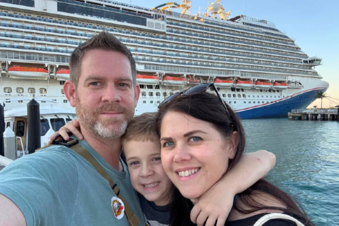 A smiling man, a young boy and a woman taking a selfie on a waterfront dock, with a large cruise ship moored behind them on calm blue water