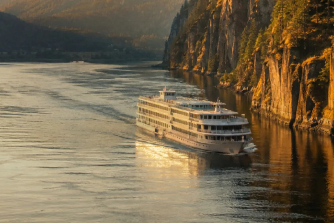 A white multi-deck river cruise ship sailing a calm river at sunset beneath tall golden cliffs and forested mountains